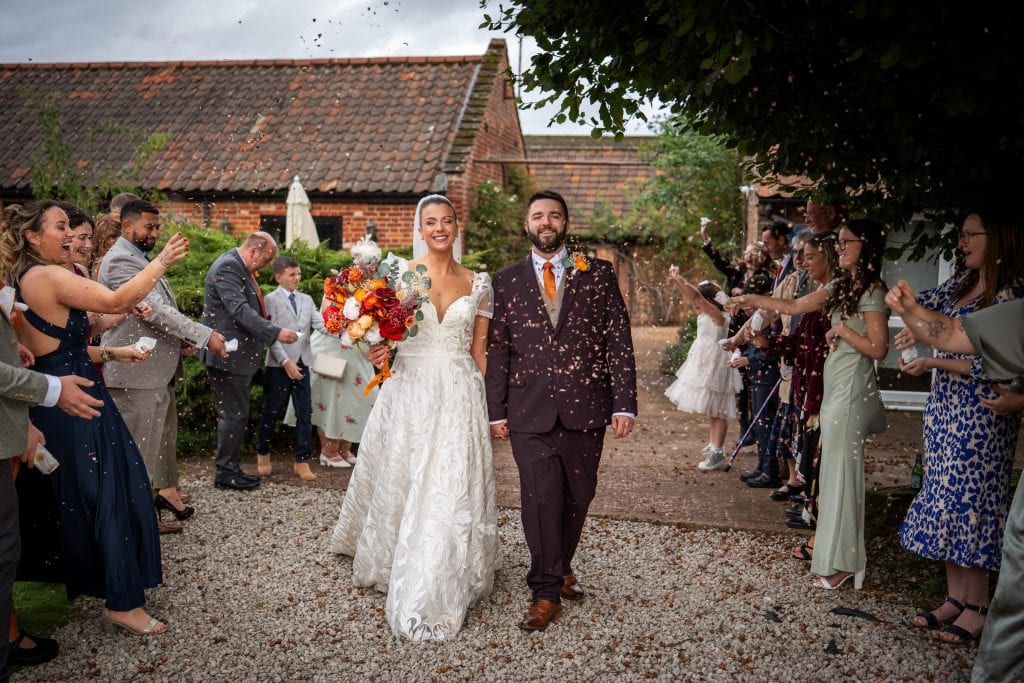 A bride and groom walk outside as guests throw confetti. The bride holds a bouquet and both are smiling, surrounded by people in formal attire near rustic buildings.