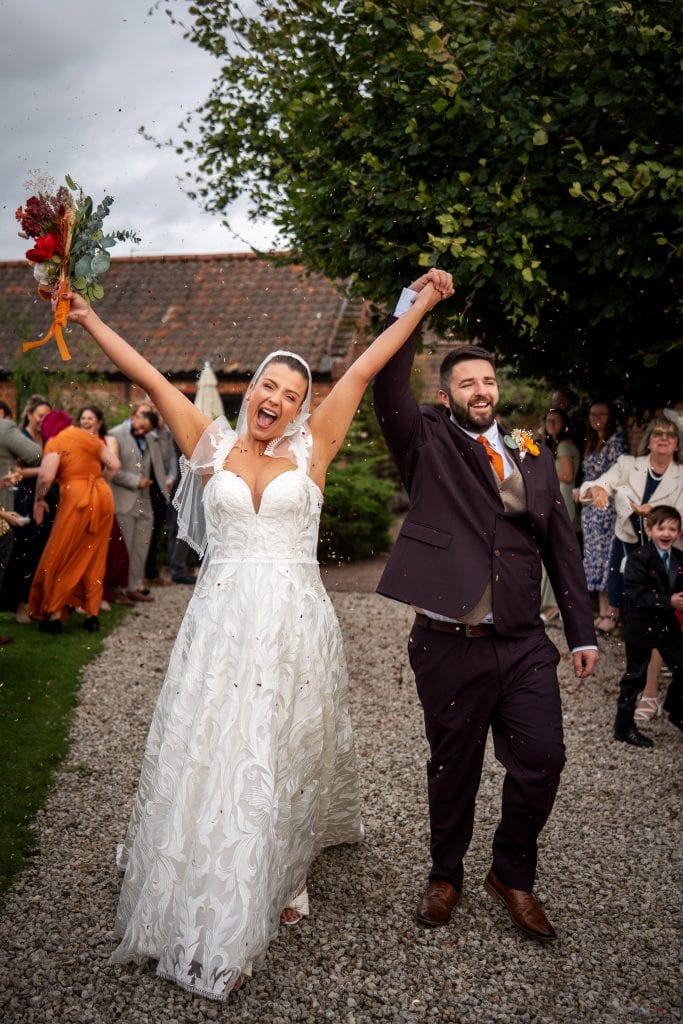 A bride and groom walk outdoors on a gravel path, holding hands and cheering, surrounded by guests at their wedding celebration.