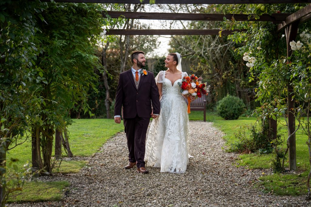 A bride and groom walk hand in hand down a garden path, surrounded by greenery, with the bride holding a colorful bouquet.