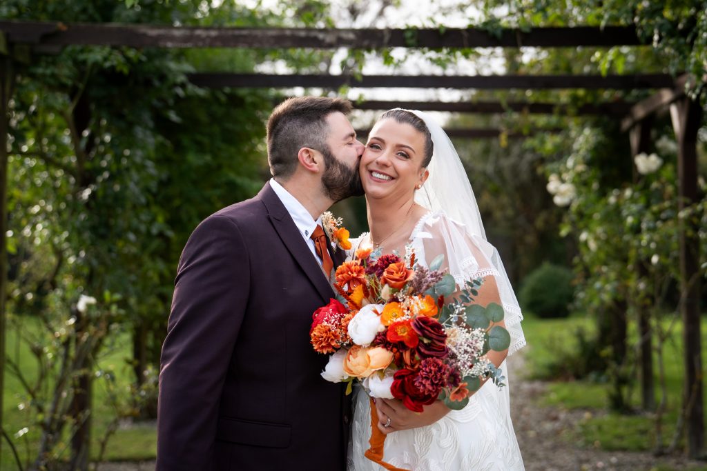 A groom in a dark suit kisses a smiling bride holding a colorful bouquet, standing together outdoors under a wooden arbor.