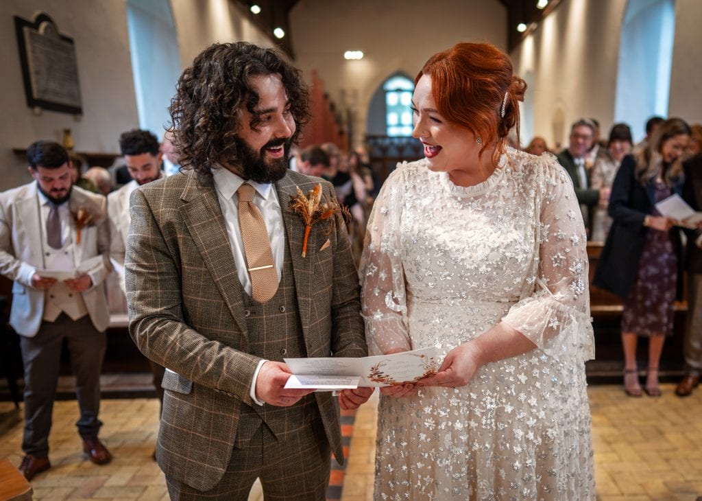 A couple in wedding attire stands together in a church, smiling and holding papers, with guests and pews visible in the background.