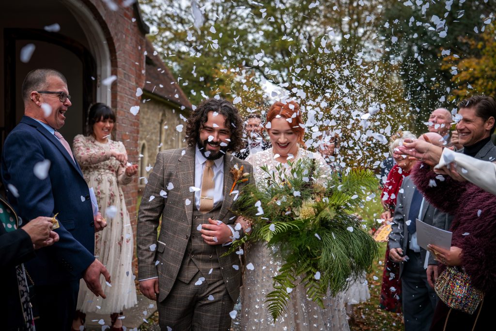 A bride and groom walk outside as guests throw confetti and celebrate; the bride holds a large bouquet and both are smiling.