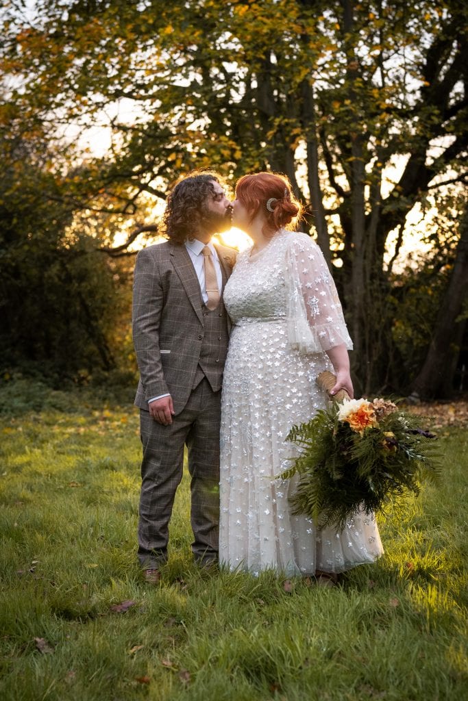 A couple in wedding attire stands on grass at sunset, kissing. The bride holds a bouquet of flowers and greenery; trees and sunlight are in the background.