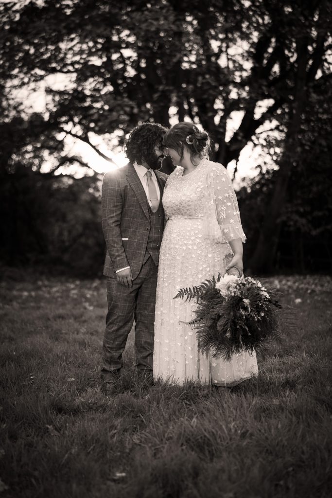 A couple stands closely in a grassy field at sunset; the person on the left wears a suit, and the person on the right wears a long dress and holds a bouquet.