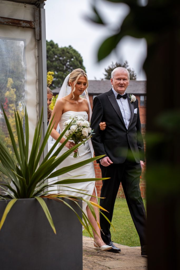 A bride in a white dress and veil, holding a bouquet, walks arm-in-arm with an older man in a suit and bow tie, outdoors near a large potted plant.