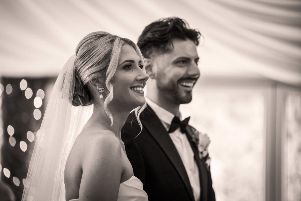 A bride and groom stand side by side indoors, smiling and looking forward. The bride wears a veil and strapless dress; the groom is in a suit with a bow tie and boutonniere.