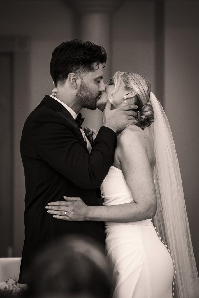 A bride and groom share a kiss indoors, dressed in wedding attire; the groom holds the bride’s face and the bride wears a veil and a strapless gown.