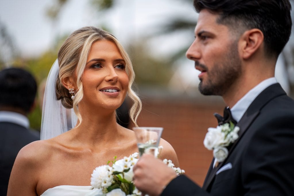 A bride holding a bouquet looks at a man in a tuxedo holding a drink, both wearing boutonnieres, standing outdoors.
