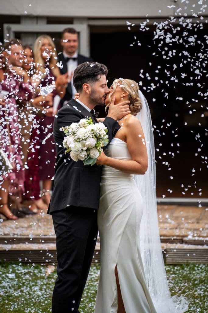 A bride and groom kiss outdoors in wedding attire as confetti falls, with guests standing and clapping in the background.