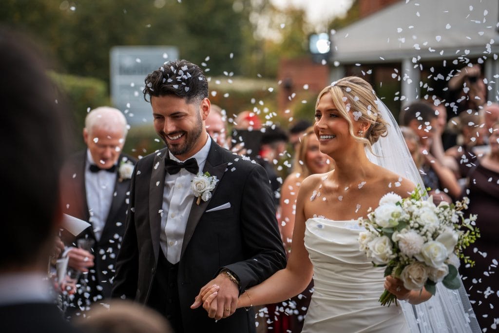 A bride and groom walk hand in hand outdoors, smiling as guests throw confetti. The bride holds a bouquet of white flowers, and both are dressed in formal wedding attire.
