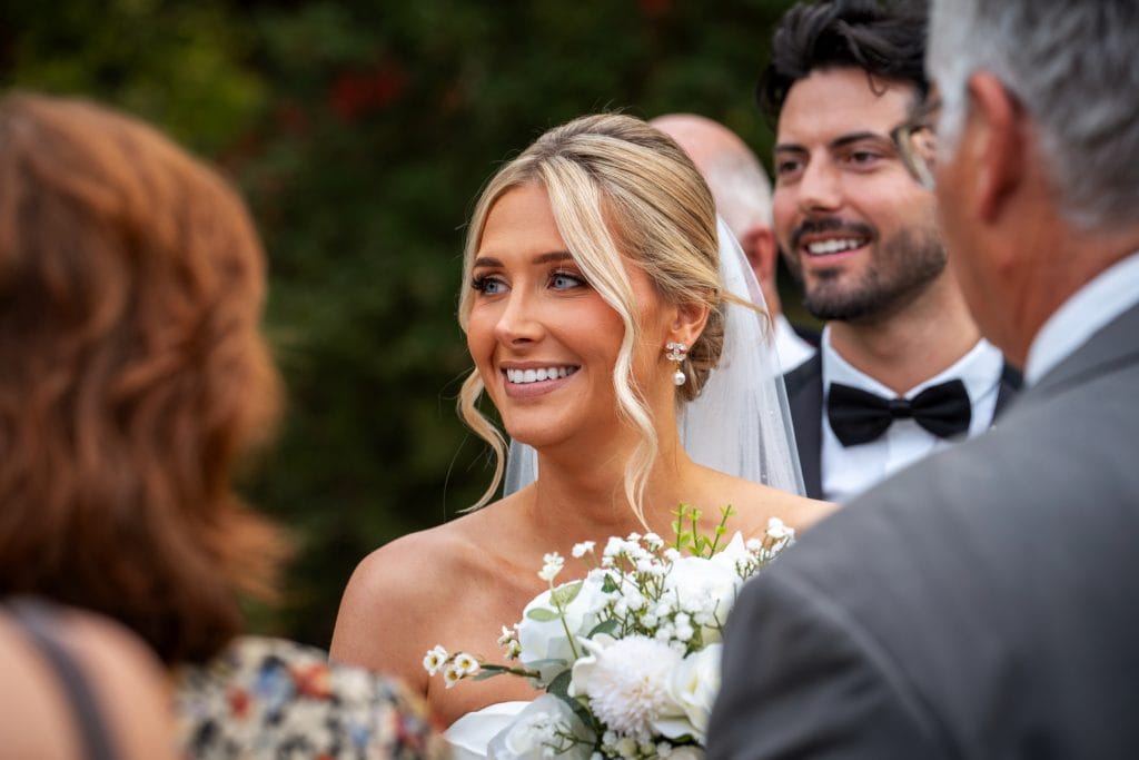 A bride in a white dress holds a bouquet of flowers and smiles among guests at an outdoor wedding ceremony.