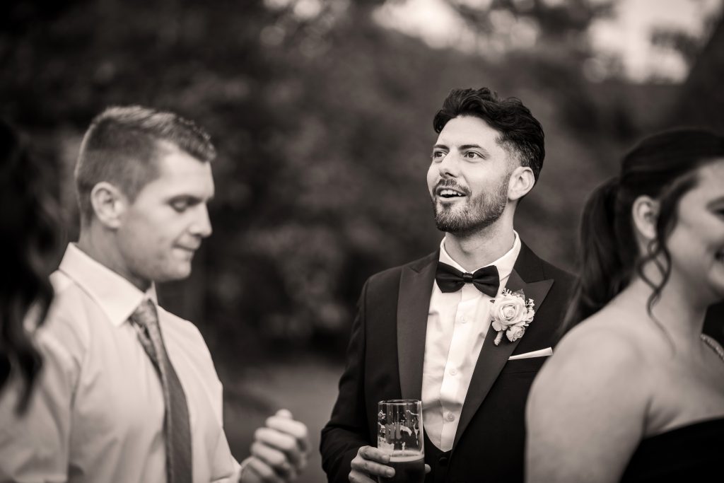 Three people are socializing outdoors; a man in a tuxedo with a boutonniere holds a drink and smiles, while two others stand nearby, one in a tie and the other in a dress.