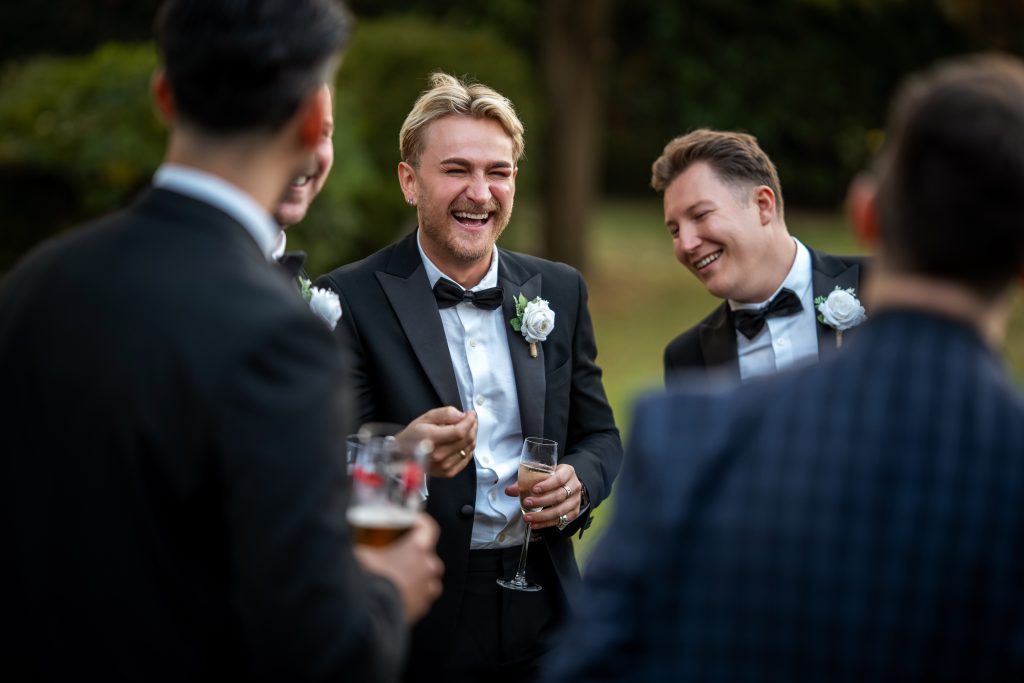 Group of men in formal suits, two holding drinks and laughing outdoors, with greenery in the background.