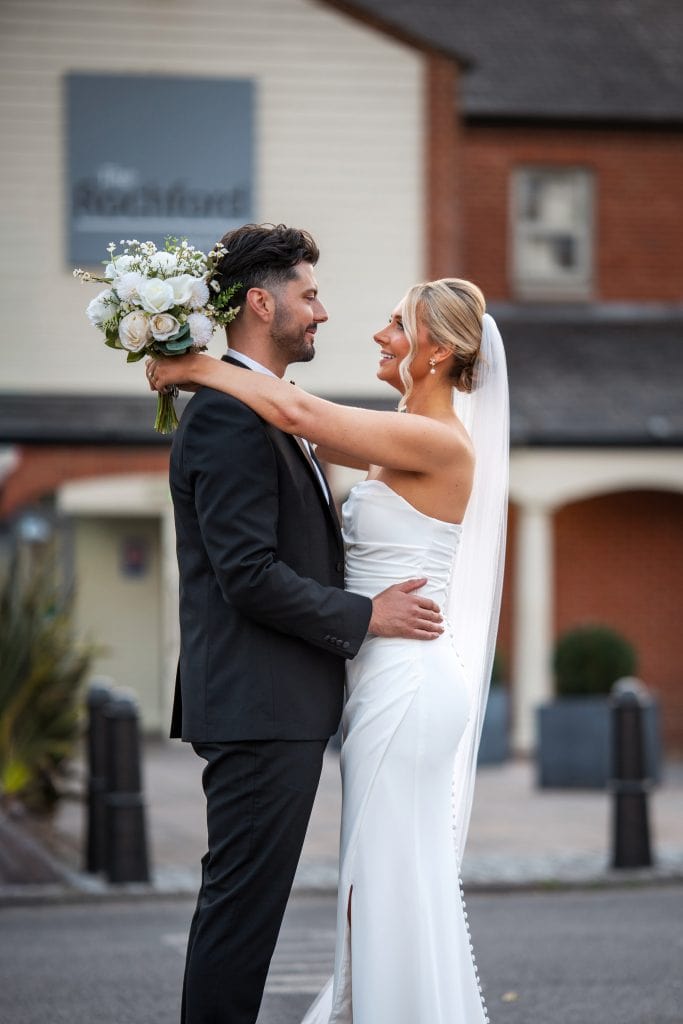 A bride in a white dress and veil holds a bouquet and embraces a groom in a black suit outdoors, both looking at each other and smiling.