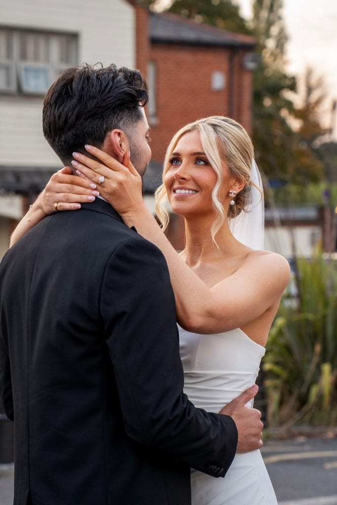 A bride in a white dress and veil holds a groom in a black suit outdoors, both smiling and looking at each other.