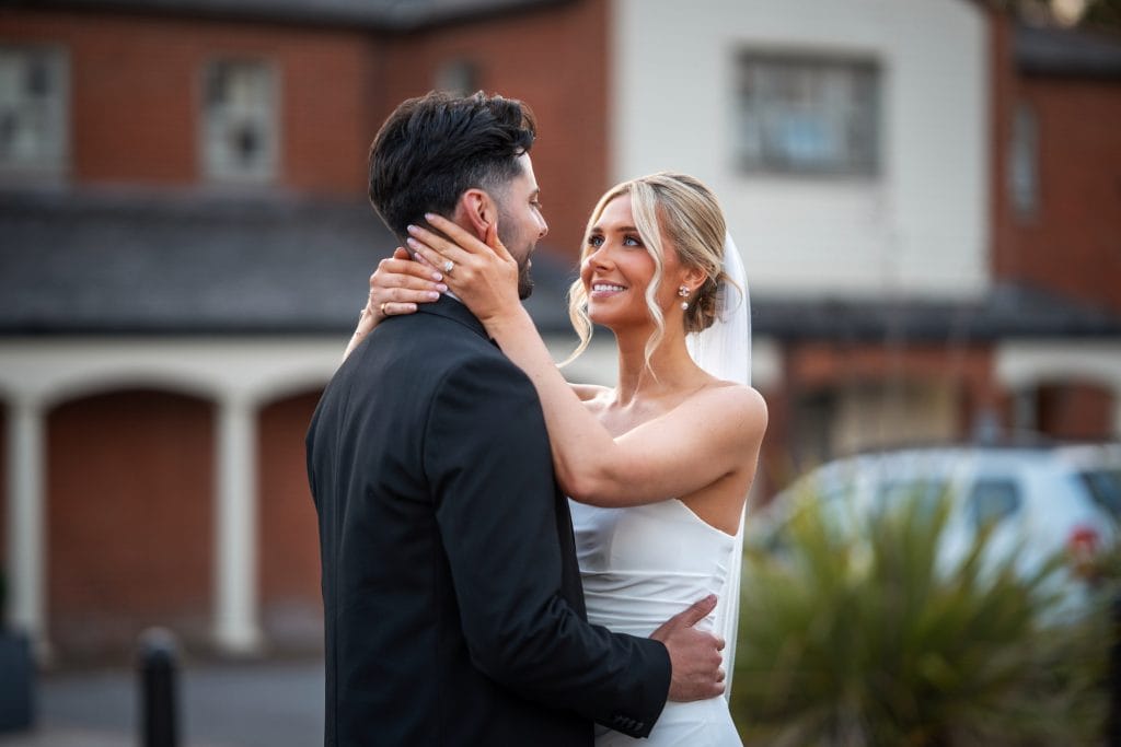 A bride in a white dress and veil holds the face of a groom in a black suit outdoors, both smiling at each other in front of a brick building.