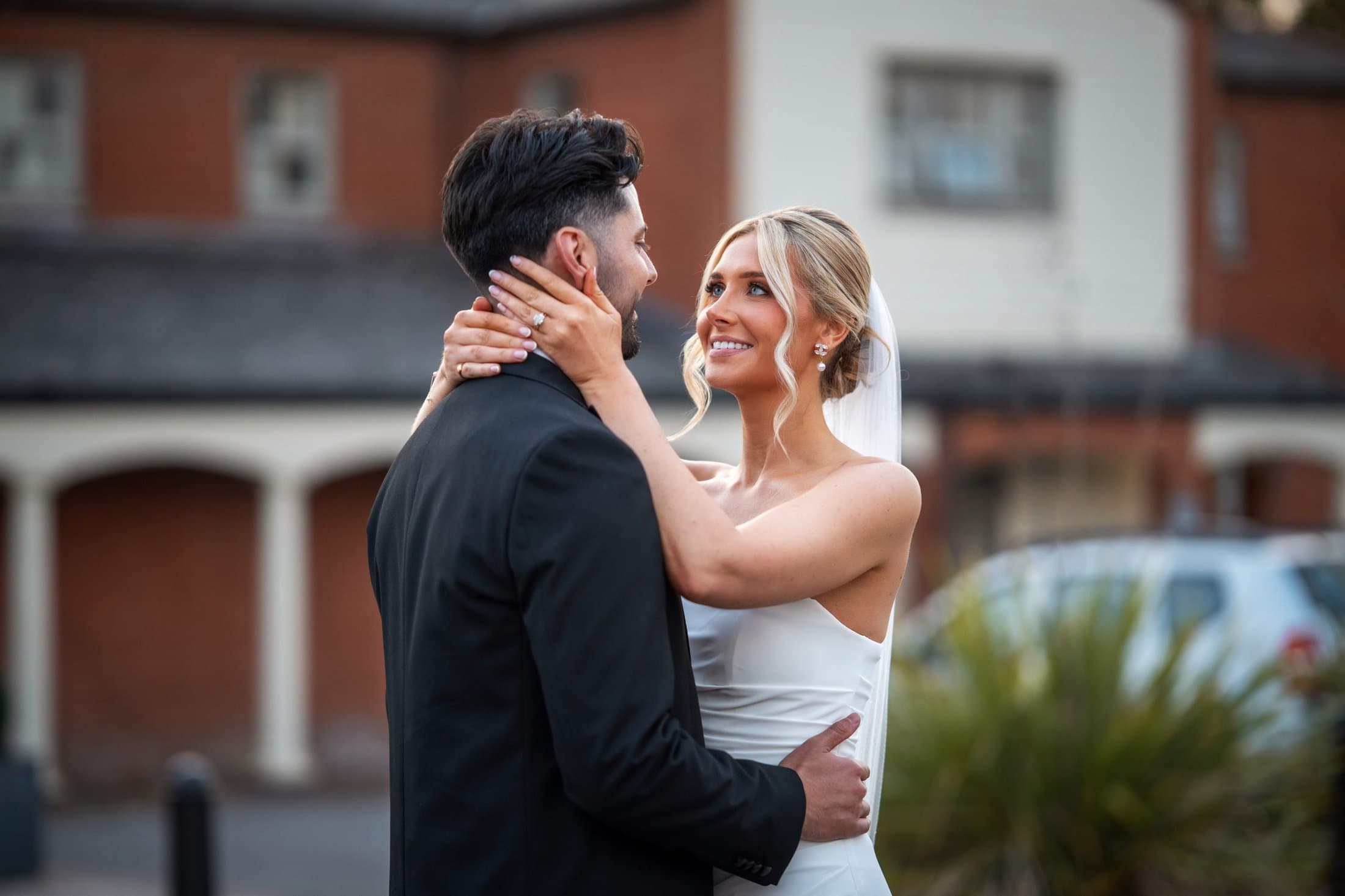 A bride in a white dress and veil holds the face of a groom in a black suit outdoors, both smiling at each other in front of a brick building.