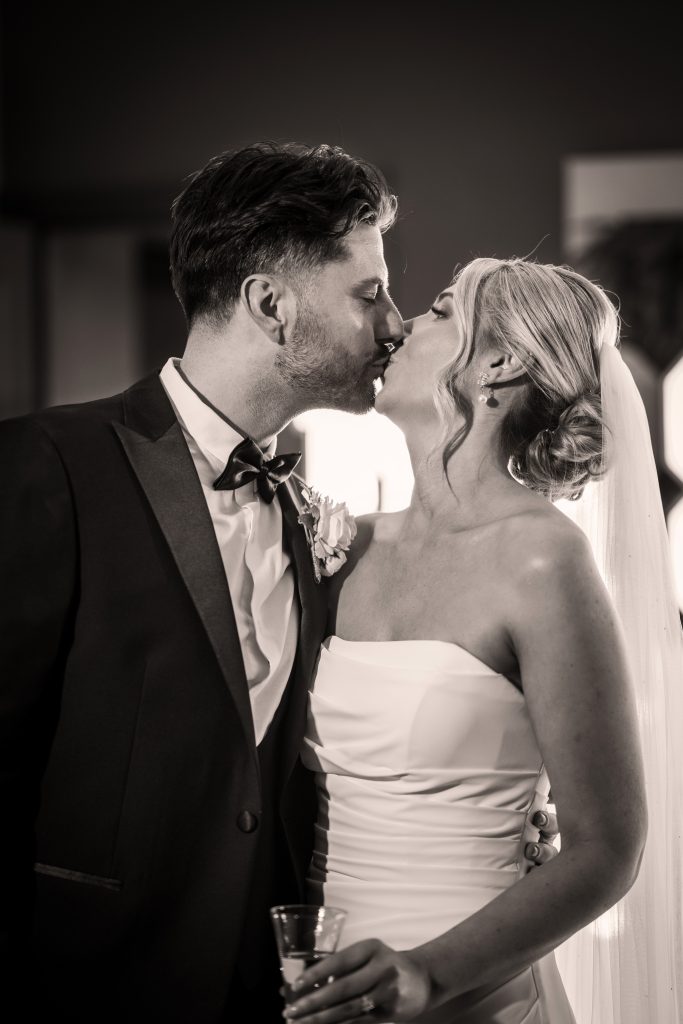 A bride and groom dressed in wedding attire share a kiss; the bride holds a drink and wears a veil, while the groom wears a suit with a bow tie.