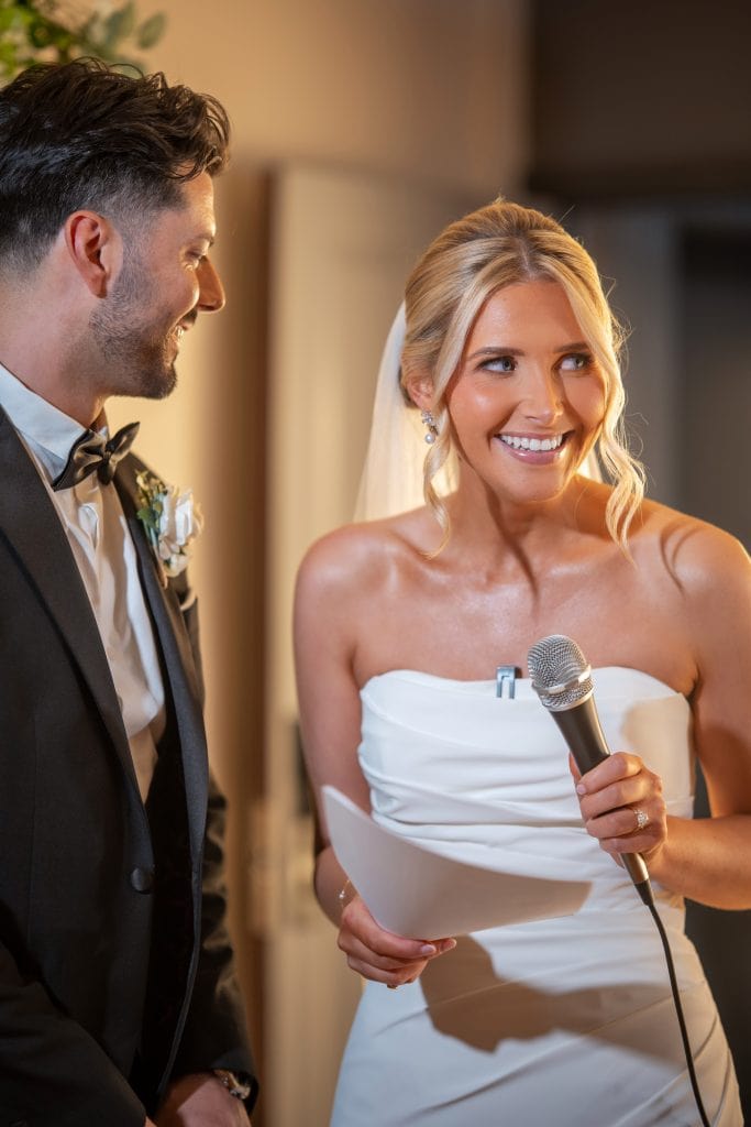 A bride in a white dress holds a microphone and paper, smiling, while a groom in a black suit stands beside her indoors.