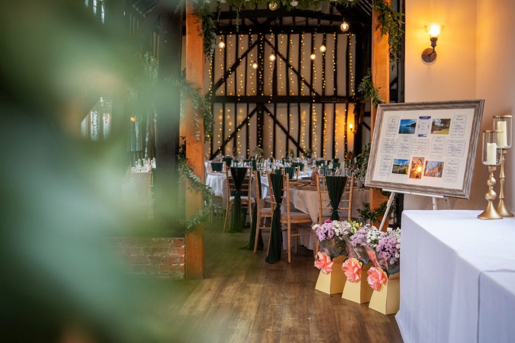 A decorated event venue with round tables, chairs, string lights, floral arrangements, framed seating chart, and candelabras, set for a formal gathering.