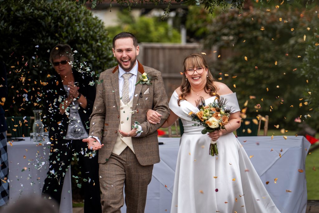 A bride and groom walk outside smiling as confetti is thrown over them. The bride holds a bouquet and wears a white dress, while the groom is in a brown checked suit.
