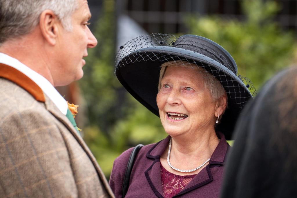 An elderly woman wearing a black hat and a pearl necklace smiles while talking to a man outdoors.