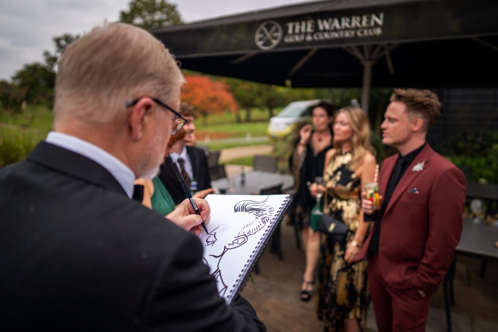 A person in a suit sketches a caricature of a man in a maroon suit at an outdoor event at The Warren Golf & Country Club, with other guests standing nearby.