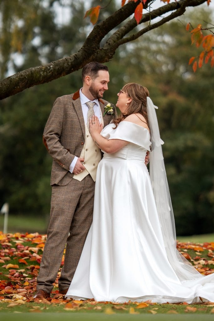 A bride and groom stand outdoors on fallen autumn leaves, facing each other and smiling, dressed in a white wedding gown and a brown plaid suit.
