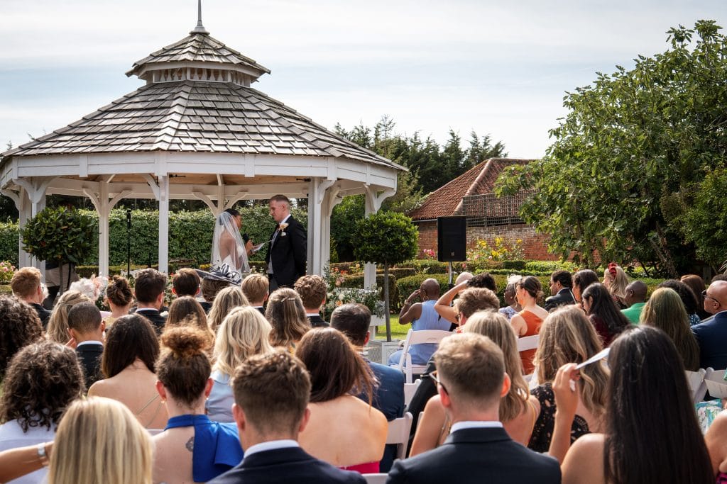 A bride and groom stand at a white gazebo exchanging vows during an outdoor wedding ceremony, with guests seated and watching. Wedding Photo taken by Jennings Photography