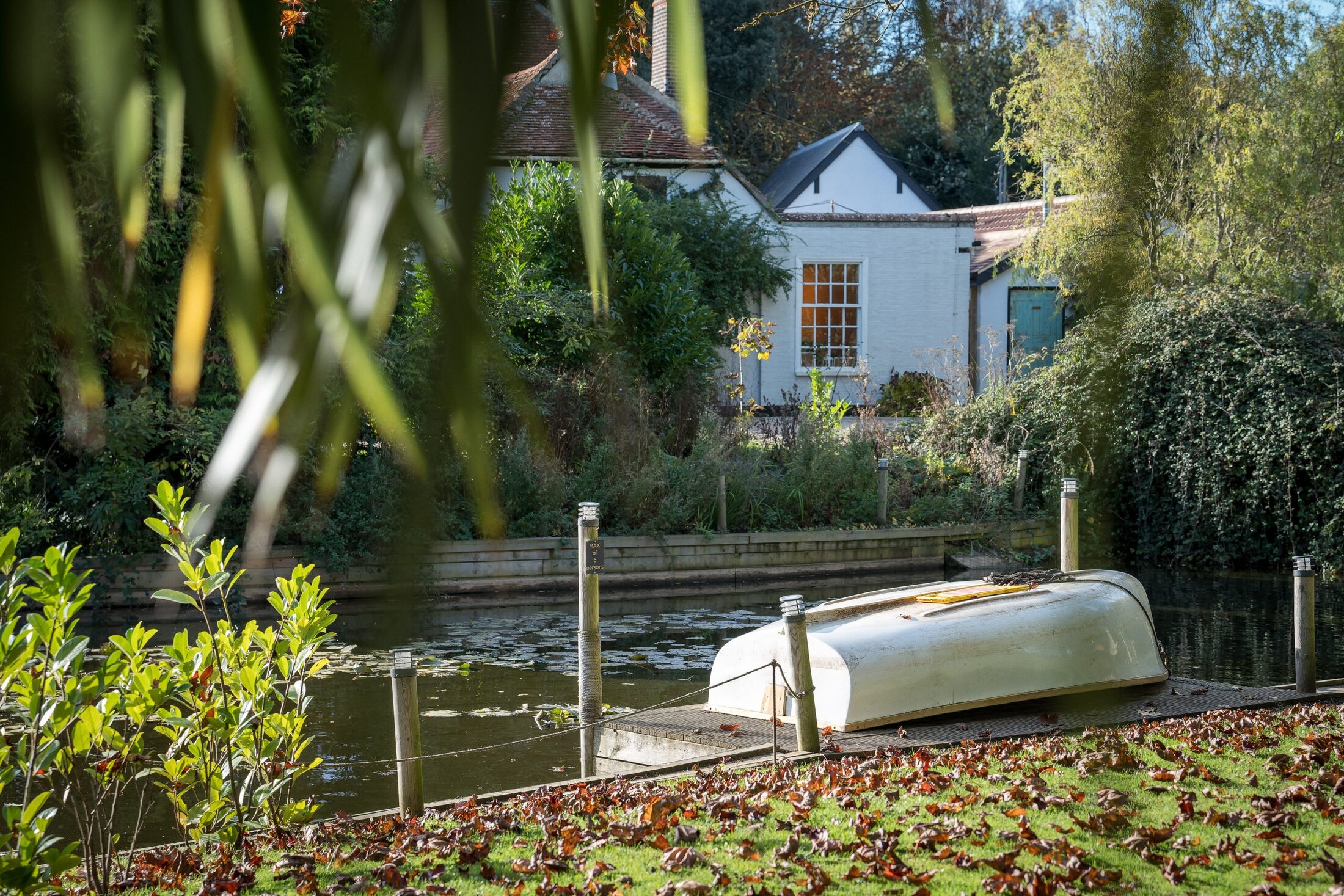 An upside-down rowboat rests on a dock by a calm river, with a white house—reminiscent of elegant Essex wedding venues—and dense greenery in the background.