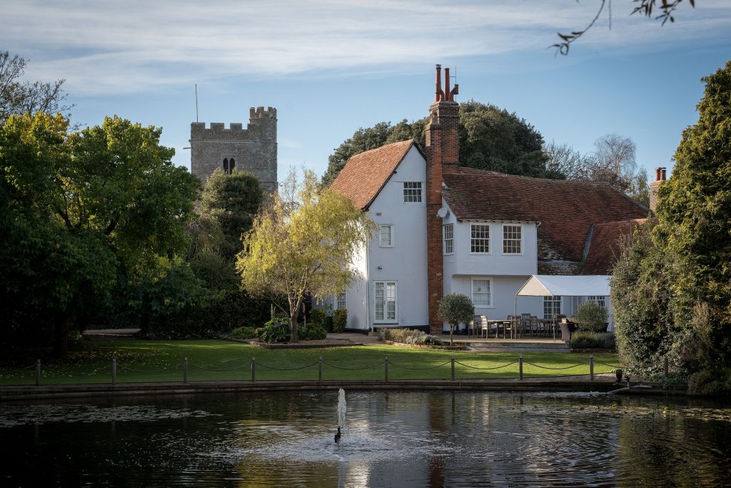 A white house with a red-tiled roof stands beside a pond, offering outdoor seating; a stone church tower rises amid trees—an idyllic scene, perfect for those seeking unique Essex wedding venues.