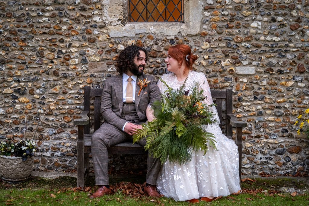 A bride and groom sit on a wooden bench outside All Saints Church, holding hands and looking at each other, with the bride holding a large bouquet of greenery.