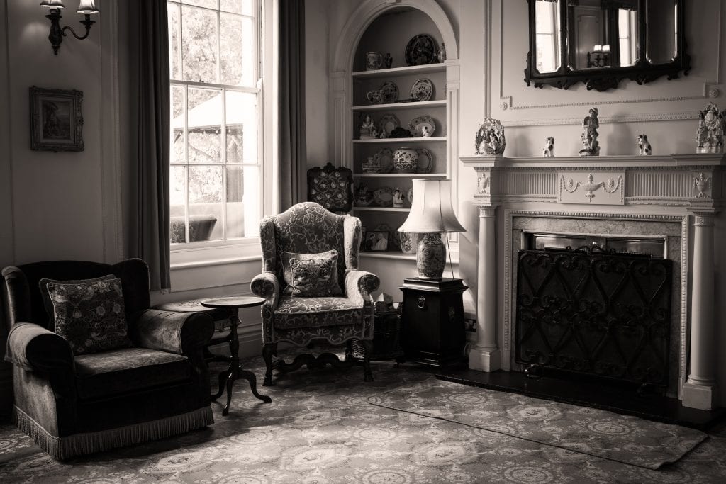 A vintage living room with ornate armchairs, a side table, shelves of decorative china, a lamp, a window, and a fireplace with figurines in black and white. Wedding Photo taken by Jennings Photography