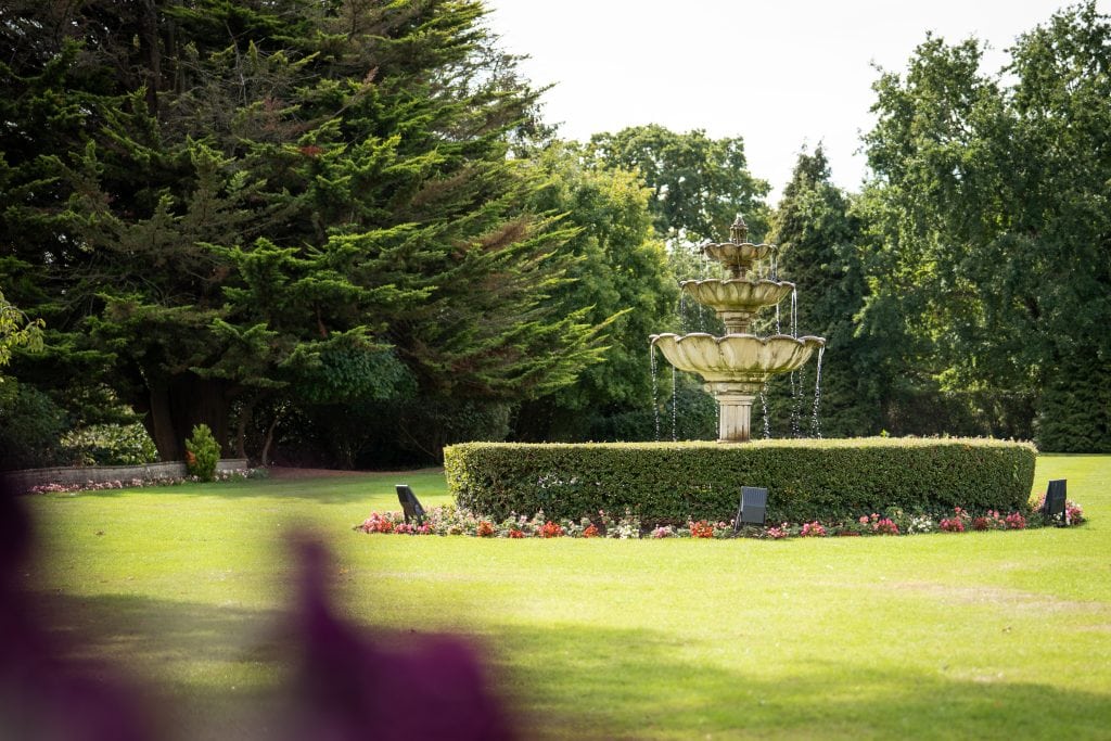 A tiered stone fountain surrounded by trimmed hedges and flower beds stands on a grassy lawn with large trees in the background. Wedding Photo taken by Jennings Photography