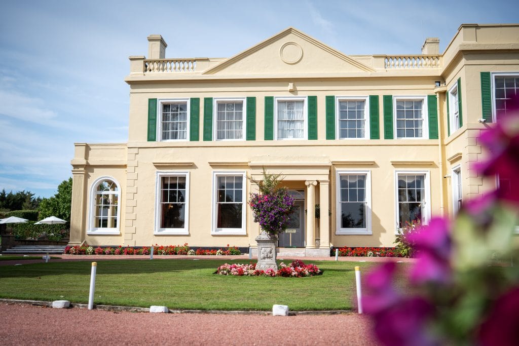 Large yellow mansion with green window shutters, a manicured lawn, colorful flower beds, and a potted plant in front; blue sky and some outdoor seating in the background. Wedding Photo taken by Jennings Photography