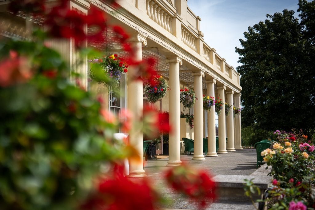 A row of cream-colored columns supports a balcony; hanging baskets with flowers decorate the colonnade, with blurred red and pink flowers in the foreground. Wedding Photo taken by Jennings Photography