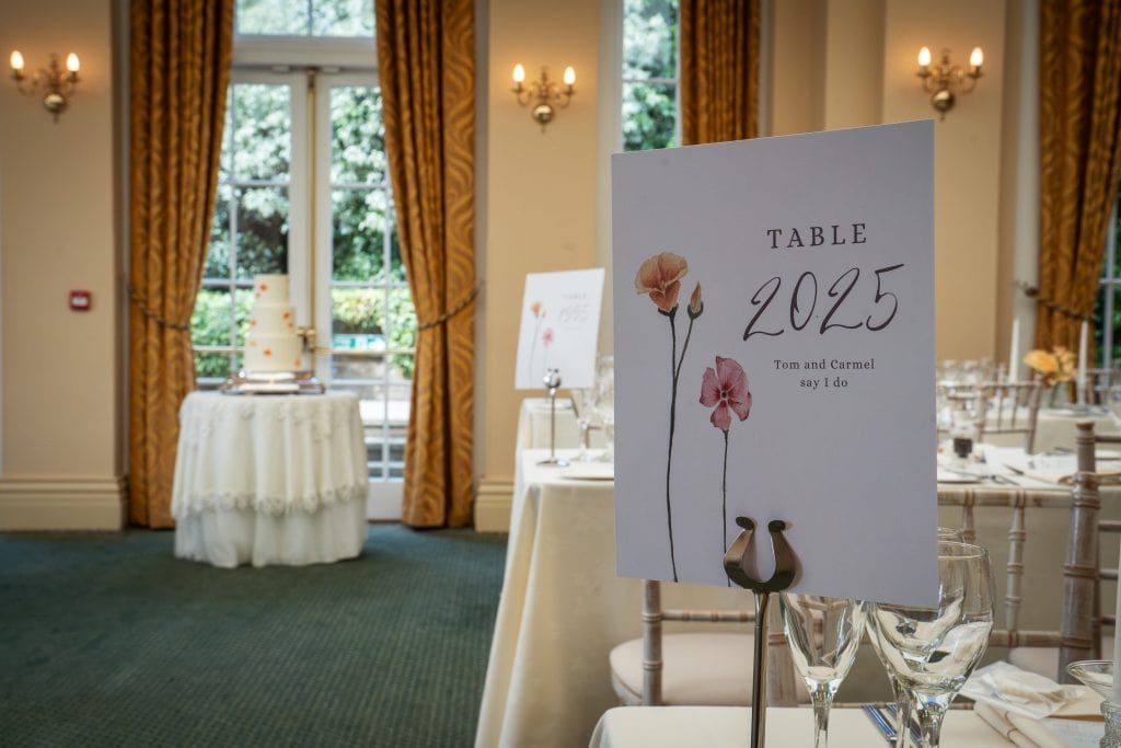 A wedding reception setup with a table card labeled "Table 2025" and a cake on a decorated table in the background near large windows with yellow curtains. Wedding Photo taken by Jennings Photography