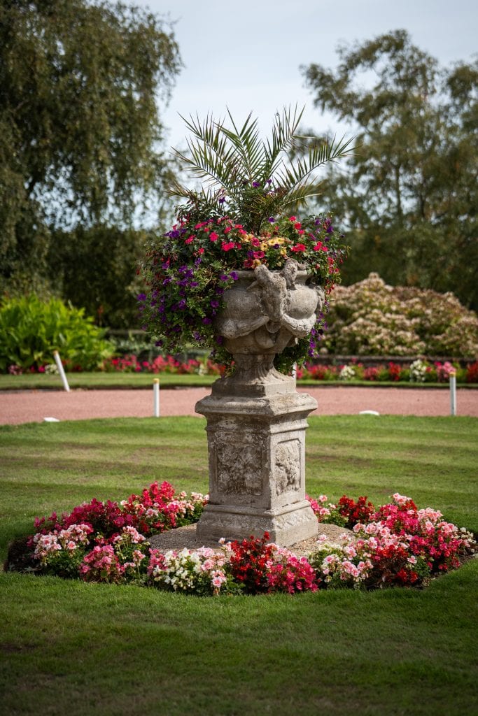 A stone pedestal planter with a palm and colorful flowers stands on a manicured lawn, surrounded by a circular flower bed in a formal garden. Wedding Photo taken by Jennings Photography