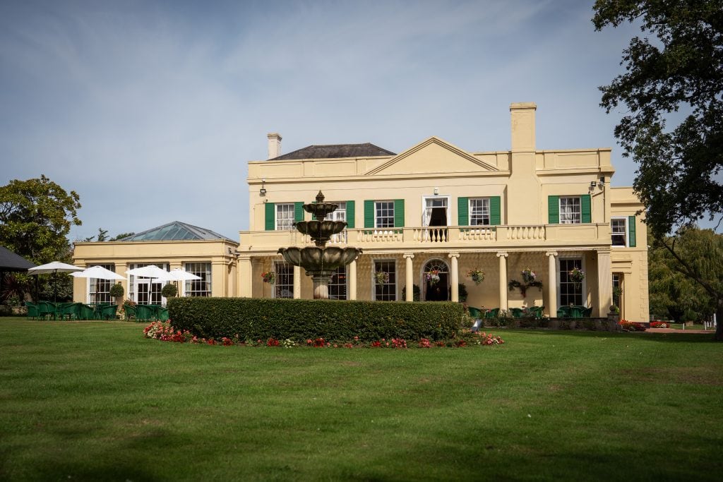 A large yellow manor house with green shutters sits behind a circular fountain and neatly trimmed lawn under a blue sky. Wedding Photo taken by Jennings Photography
