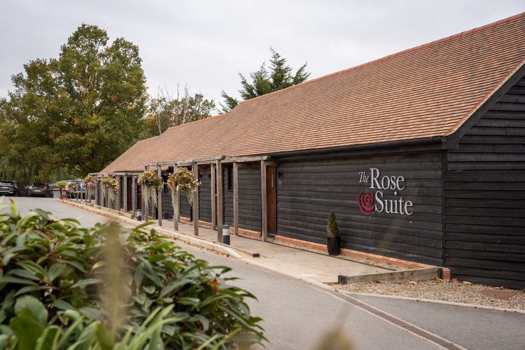 A long, dark wooden building with a red-tiled roof and "The Rose Suite" sign on the exterior, surrounded by plants and a parking area. Wedding Photo taken by Jennings Photography