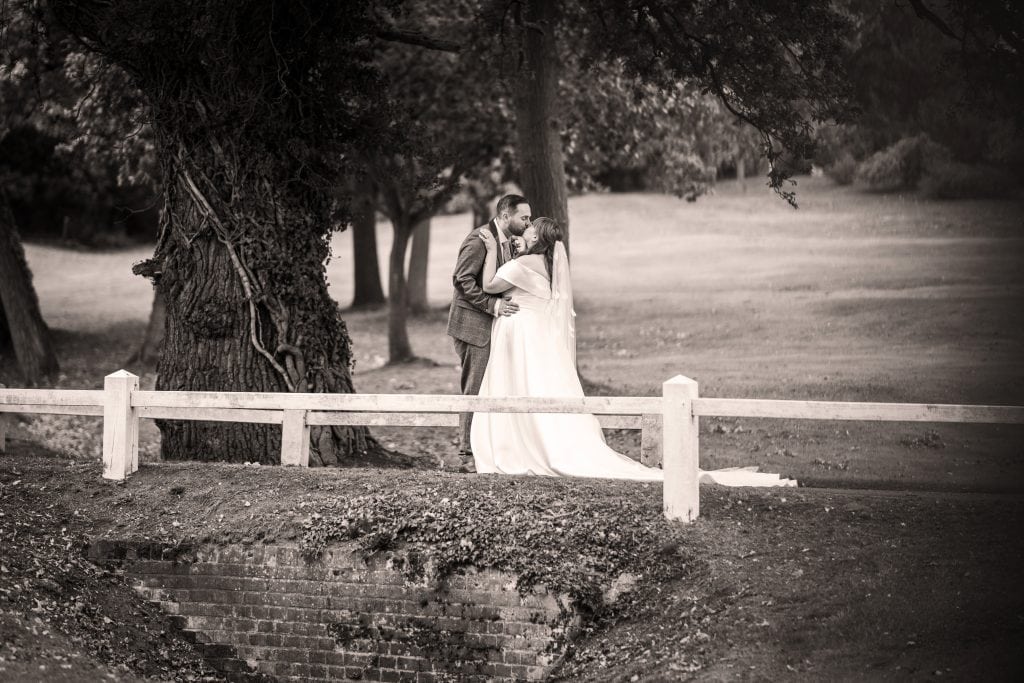A bride and groom stand outdoors by a wooden fence, embracing under trees, with grass and a brick-lined ditch in the foreground. Wedding Photo taken by Jennings Photography