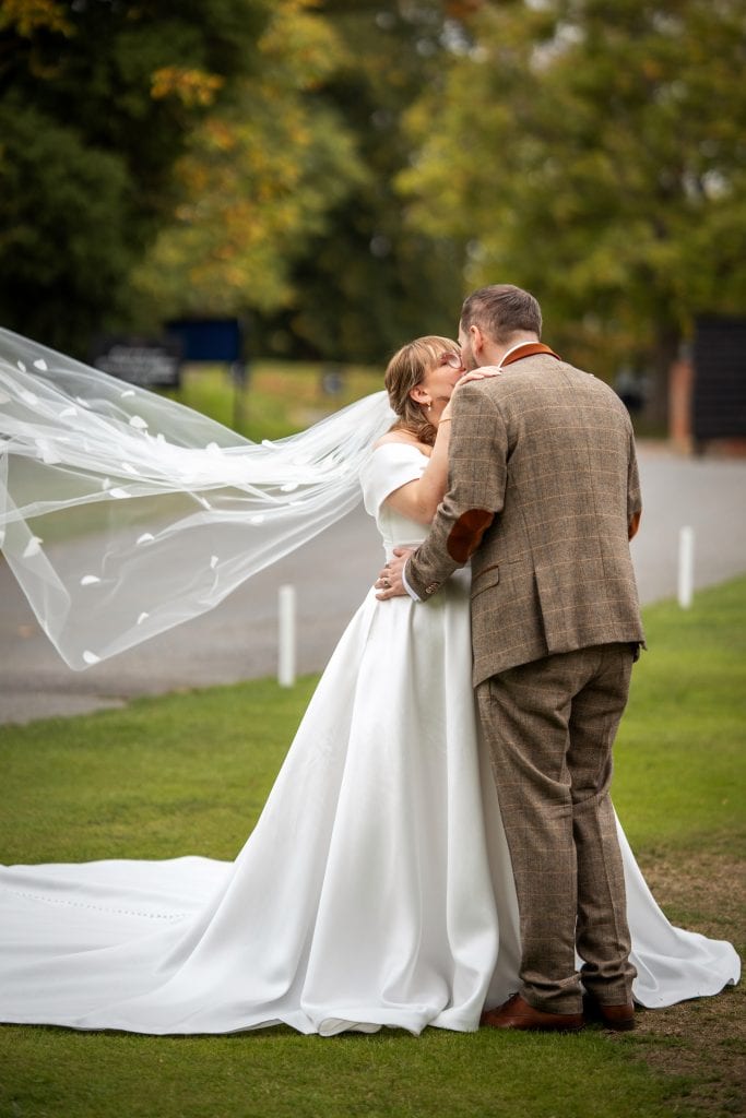 A bride and groom kiss outdoors; the bride's veil is flowing in the wind and they are standing on grass with trees in the background. Wedding Photo taken by Jennings Photography