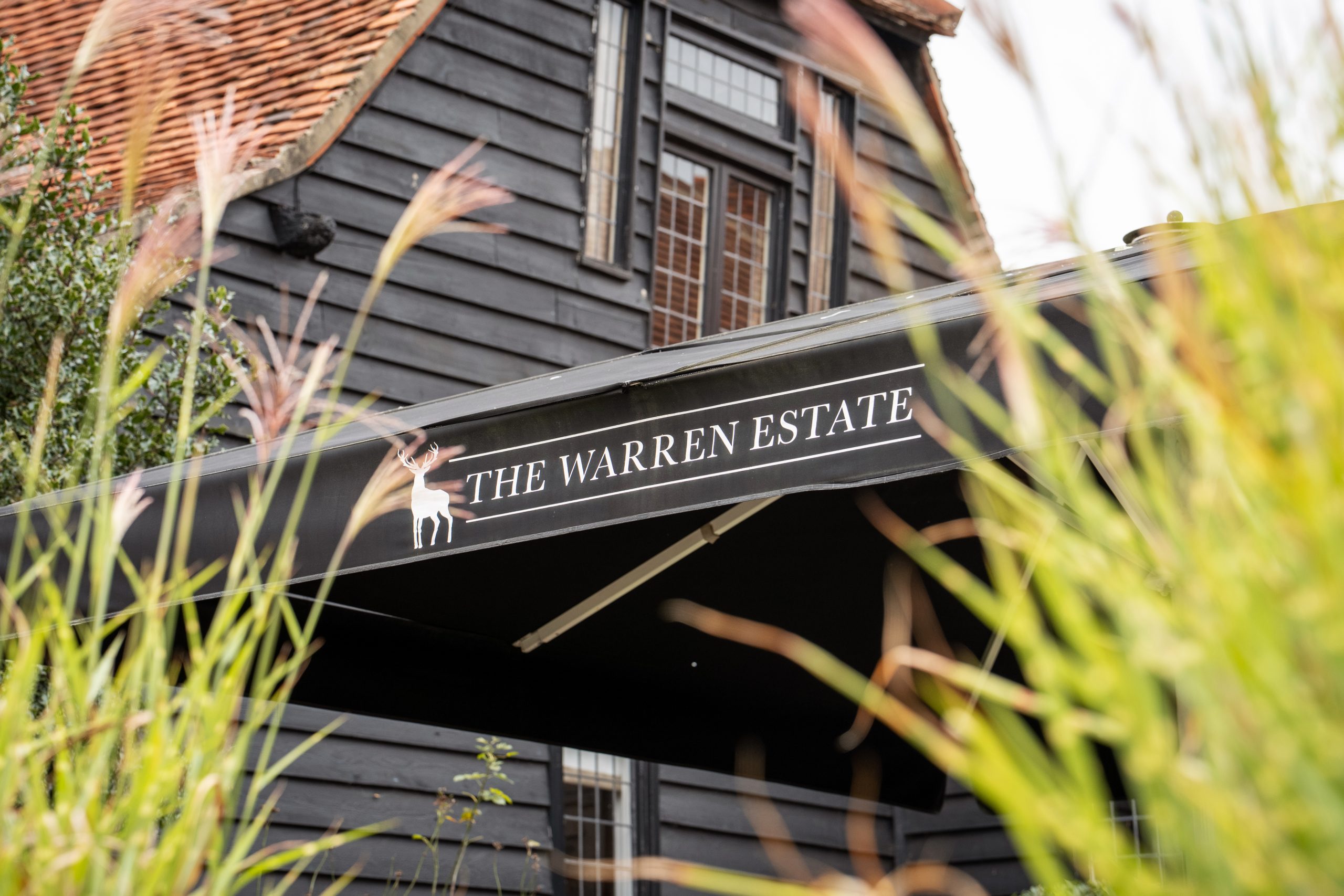 A black awning with "The Warren Estate" text and a deer logo in front of a dark wooden building, partially obscured by tall grass. Wedding Photo taken by Jennings Photography