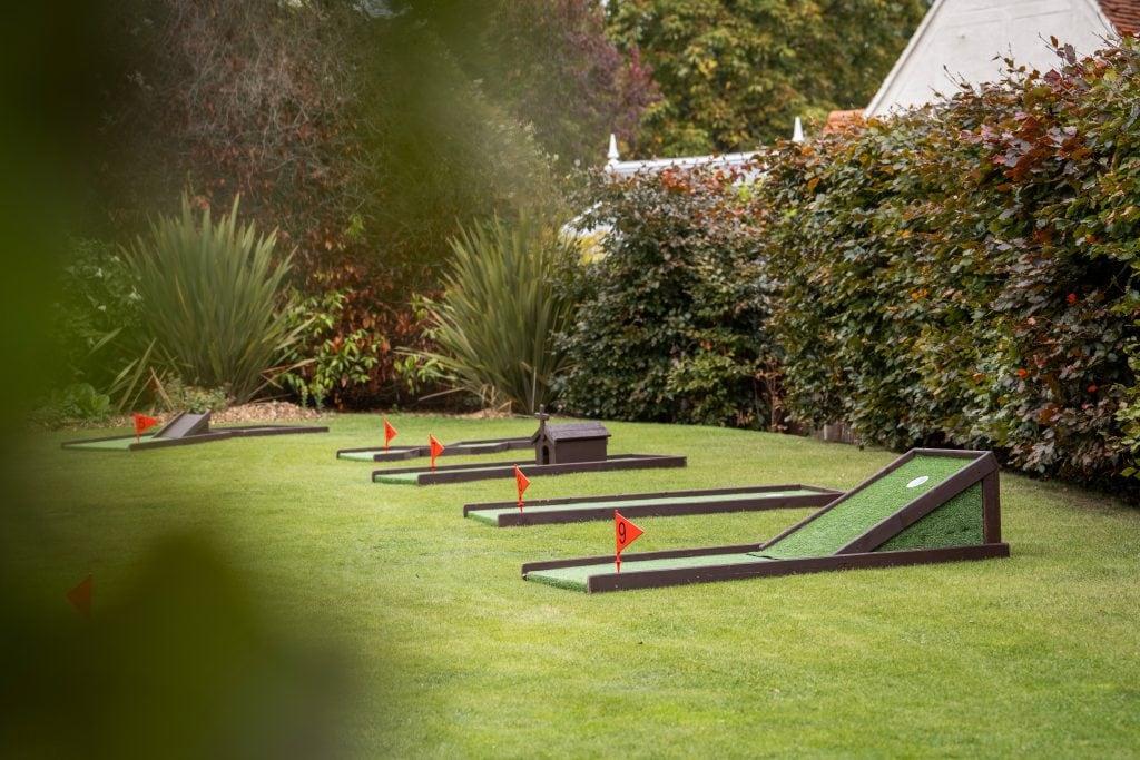 Mini-golf course with several obstacles and red flags set up on a grassy lawn, surrounded by bushes and trees. Wedding Photo taken by Jennings Photography