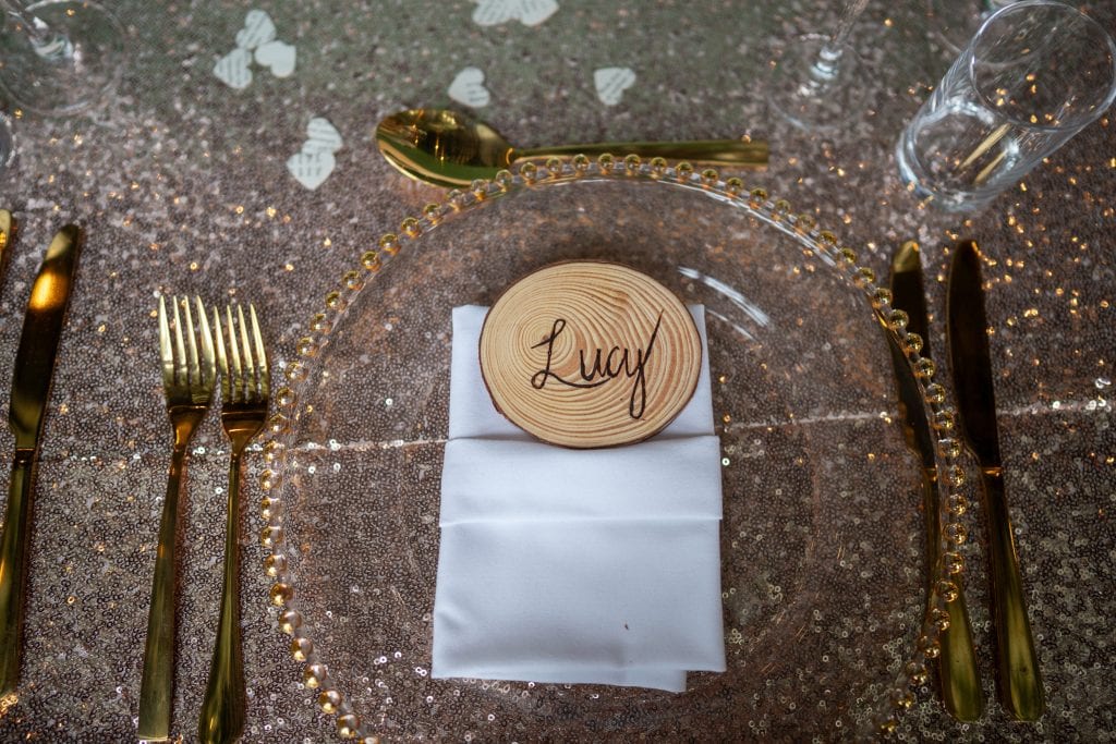 A clear glass plate with gold beads sits on a glittery tablecloth, holding a folded white napkin and a wooden place card labeled "Lucy," surrounded by gold cutlery and glassware. Wedding Photo taken by Jennings Photography
