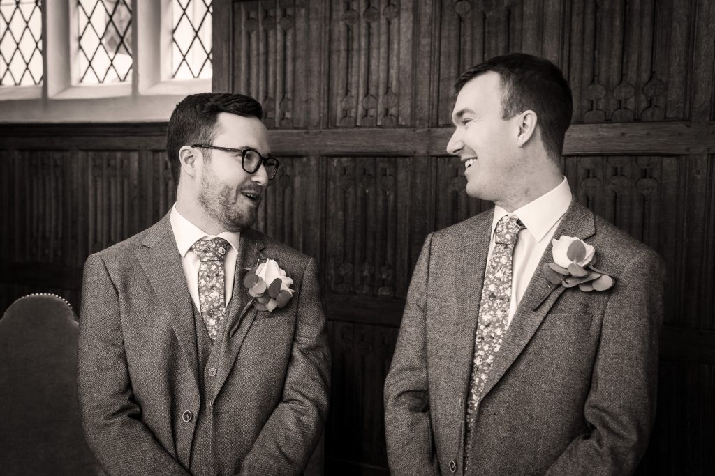 Two men in suits with floral ties and boutonnieres stand indoors, facing each other and smiling, in front of a wooden paneled wall and leaded glass windows. Wedding Photo taken by Jennings Photography
