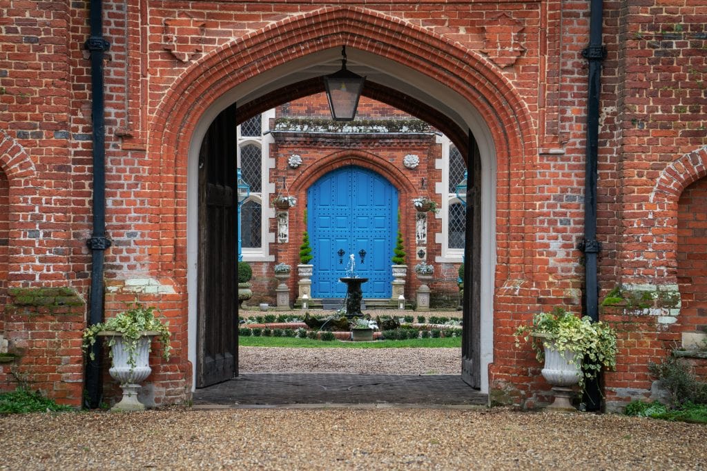 A brick archway frames a courtyard with a blue double door, fountain, decorative plants, and gravel pathway. Wedding Photo taken by Jennings Photography
