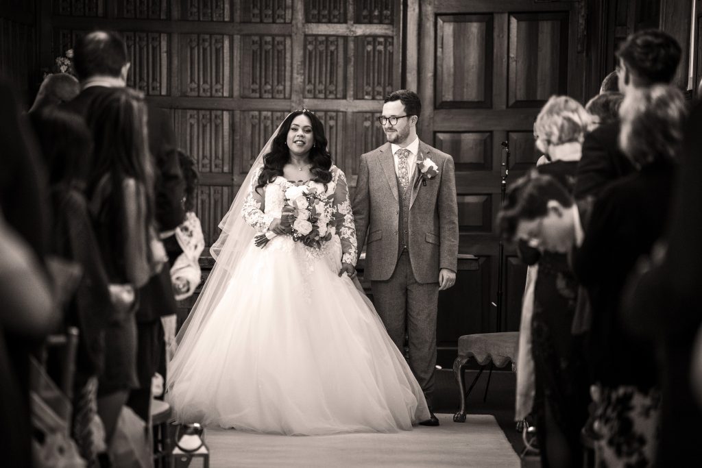 A bride in a white gown and groom in a suit walk down the aisle together, surrounded by seated guests in a wood-paneled room. Wedding Photo taken by Jennings Photography