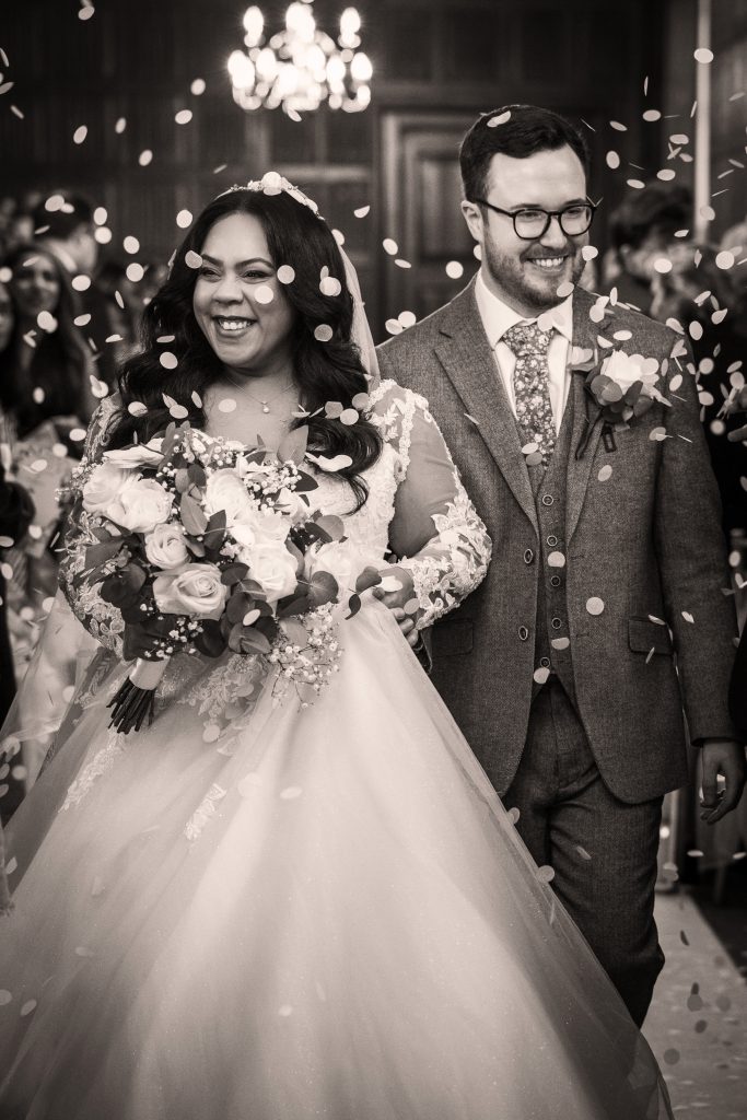 A bride and groom smile as they walk down the aisle together, surrounded by guests throwing confetti. The bride holds a bouquet of flowers. Wedding Photo taken by Jennings Photography