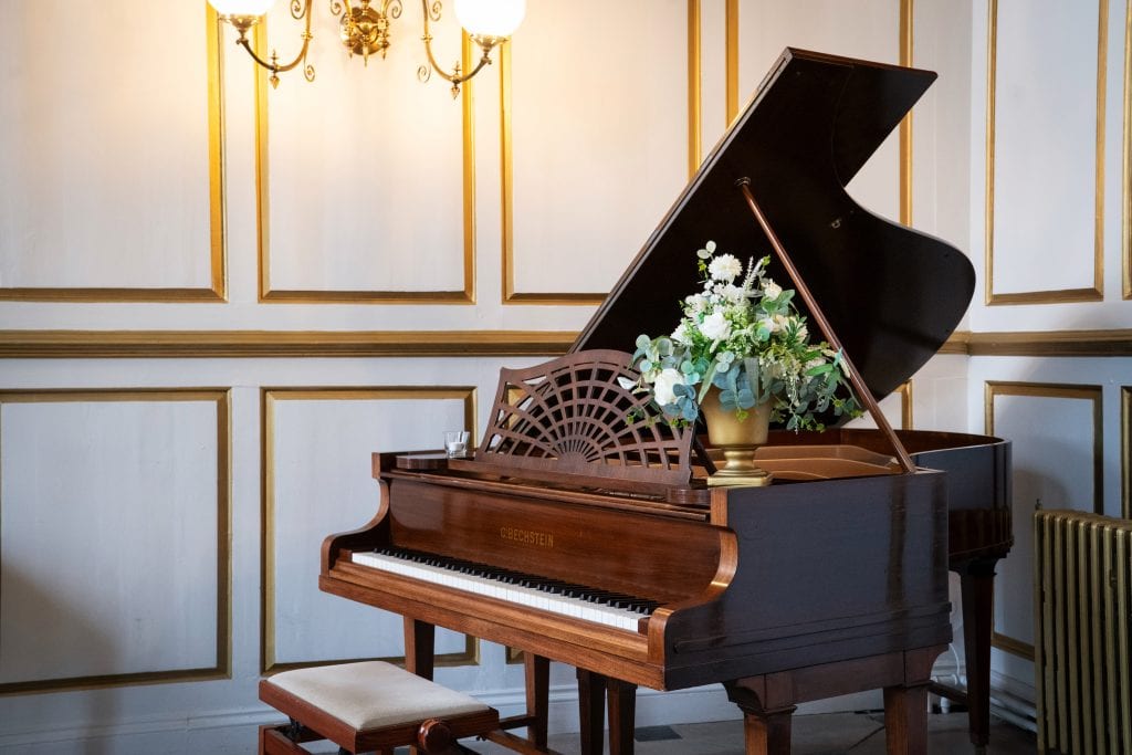A grand piano with a floral arrangement sits in an elegant room with white and gold paneled walls and a lit wall sconce. Wedding Photo taken by Jennings Photography